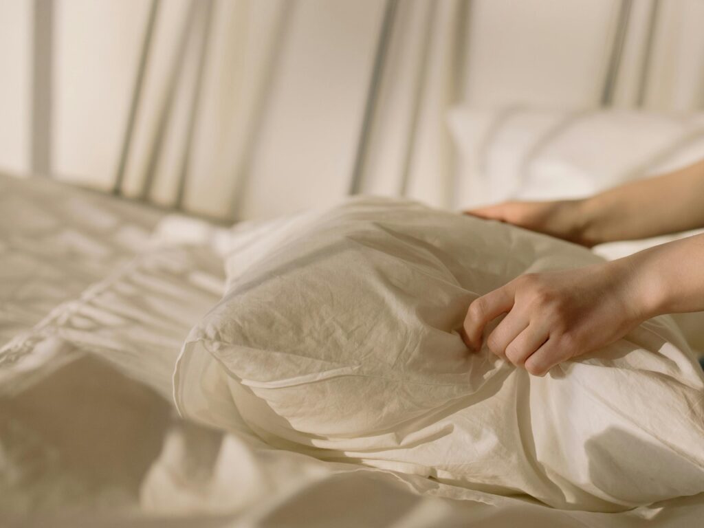 Hands arranging a pillow on a bed with light beige linens, creating a cozy bedroom ambiance.